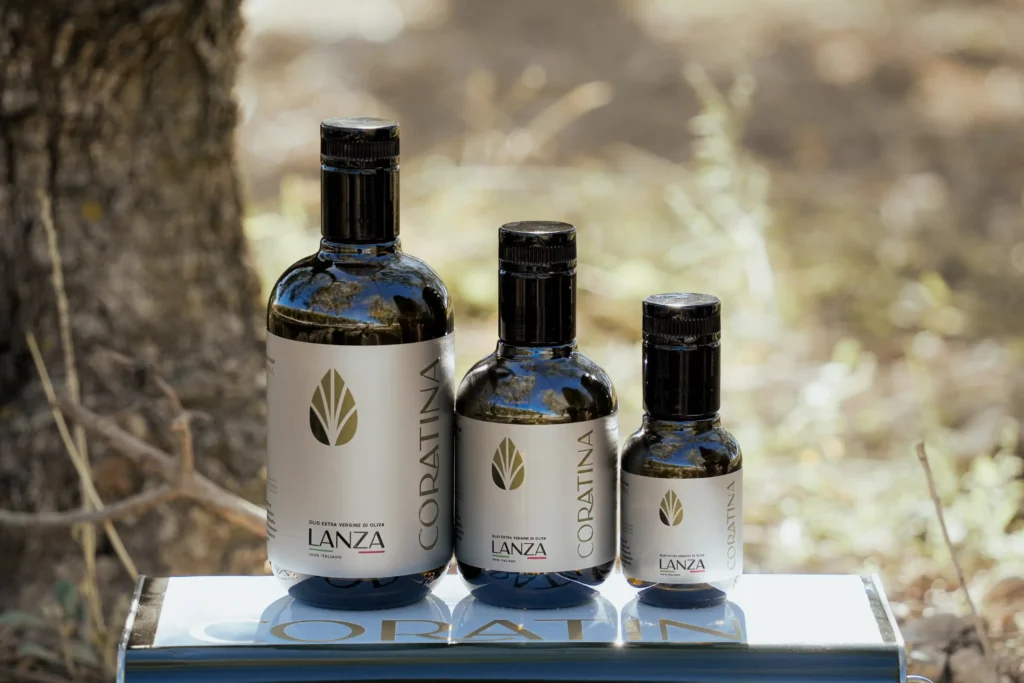 Bottiglie di olio extravergine d’oliva Coratina Lanza disposte frontalmente su scatola, sfondo rurale di uliveto a Cerignola in Puglia